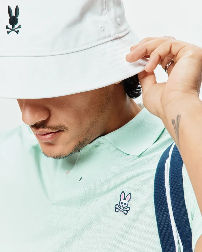 Mens Classic Pique Polo in pale mint green pique, classic fit, with an embroidered pink, black, white Psycho Bunny logo on the left chest and a navy/white striped sleeve. White bucket hat with black Psycho Bunny logo.