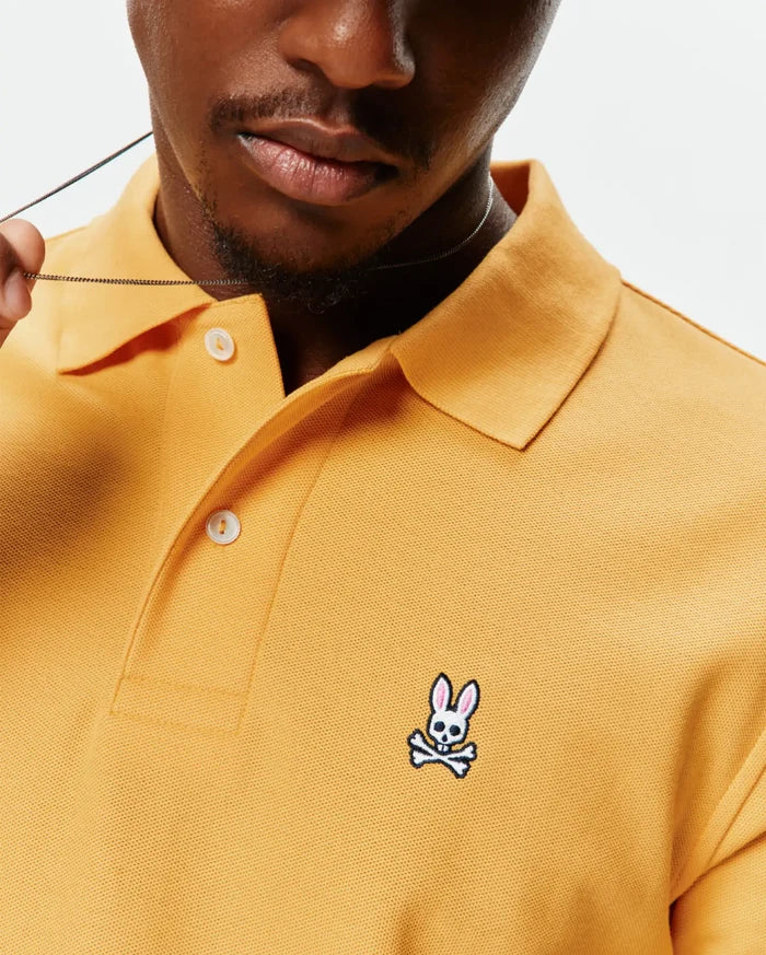 Mens Classic Pique Polo in mustard yellow, featuring a classic fit, textured pique fabric, white button placket, and a small embroidered white and pink Psycho Bunny skull logo on the left chest.