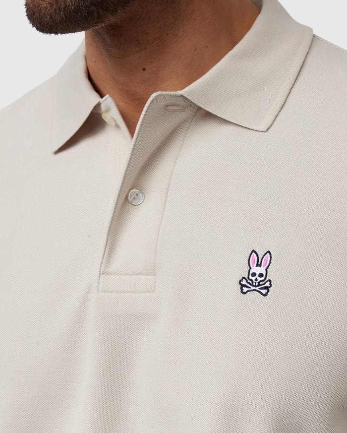 Close-up shot of a person wearing a light beige Psycho Bunny MENS CLASSIC PIQUE POLO SHIRT - B6K001B200 with a collar and buttons. This timeless shirt, crafted from luxurious Pima cotton, features an embroidered logo on the left chest area depicting a pink bunny head above crossed bones. Only part of the person’s face and neck are visible.