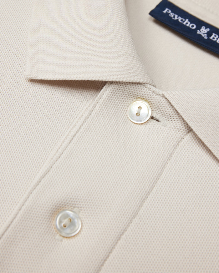 Close-up of a beige Psycho Bunny MENS CLASSIC PIQUE POLO SHIRT - B6K001B200 collar with two white buttons. The timeless shirt fabric has a textured, waffle-like pattern. A navy blue label with white text is partially visible near the collar, hinting at its luxurious Pima cotton composition.