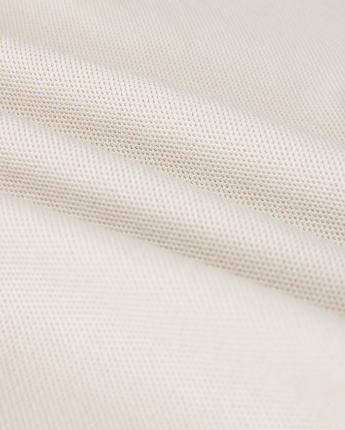 Close-up of a softly folded, light beige textured fabric with a subtle grid pattern, showcasing its smooth and pliable nature. The gentle folds add depth and highlight the luxurious Pima cotton's fine weave and soft sheen, hinting at the perfect fit for a Psycho Bunny Men's Classic Pique Polo Shirt - B6K001B200.