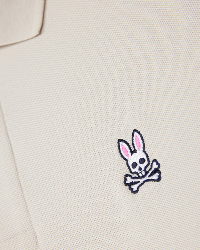 Close-up of a beige textured fabric from the Psycho Bunny MENS CLASSIC PIQUE POLO SHIRT - B6K001B200, with an embroidered logo featuring a white skull and crossbones with pink-eared bunny ears on the chest area. Crafted from luxurious Pima cotton for a perfect fit.