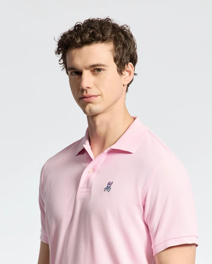 A person with curly hair wears a pale pink Psycho Bunny MENS CLASSIC PIQUE POLO SHIRT - B6K001CRPC, featuring a small embroidered logo on the chest, against a neutral background. The Pima cotton adds comfort as they look toward the camera with a slight smile.