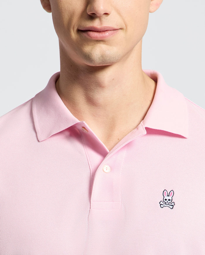 A person wears the Psycho Bunny MENS CLASSIC PIQUE POLO SHIRT - B6K001CRPC in light pink, featuring a collared, button design. Made from soft Pima cotton, it showcases an embroidered rabbit above crossbones logo on the chest against a plain light gray background.