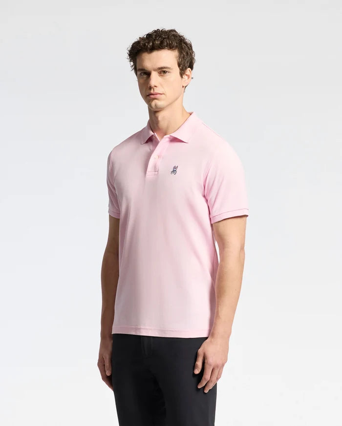 A man in a Psycho Bunny MENS CLASSIC PIQUE POLO SHIRT - B6K001CRPC in light pink, featuring a small dark logo on the chest, pairs it with dark pants against a plain light background. Crafted from luxurious Pima cotton, this polo speaks of style and elegance.
