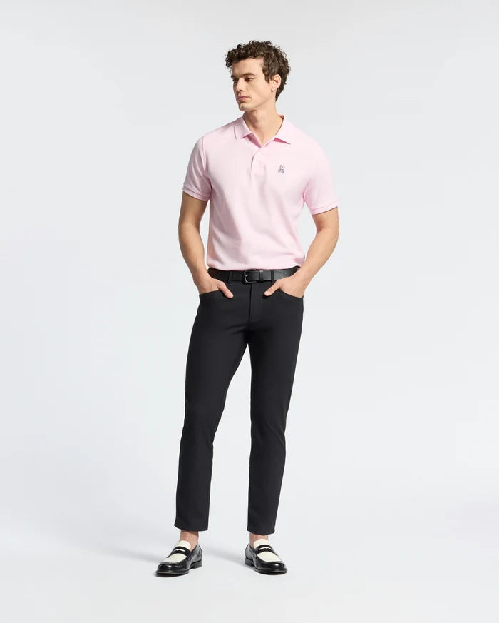 A person stands wearing a light pink Psycho Bunny MENS CLASSIC PIQUE POLO SHIRT - B6K001CRPC and black pants, hands in pockets. Two-tone loafers complete the look against a white background, highlighting the Pima cotton clothing.