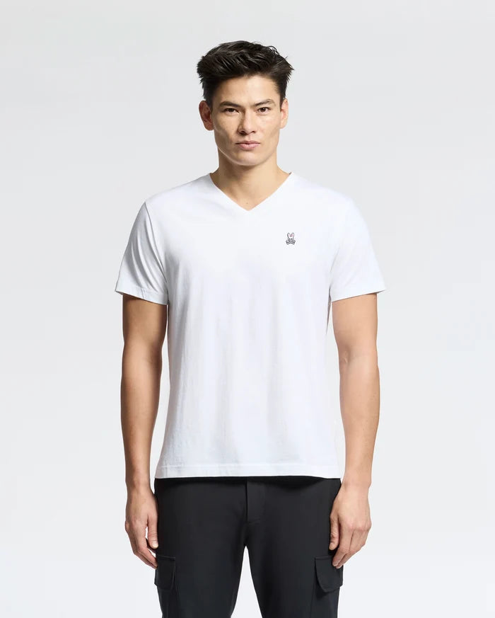 Against a light background, a person wears the Psycho Bunny Men's Classic V-Neck Tee in white with an embroidered logo, paired with black pants. The 100% Pima cotton tee complements their short dark hair as they look directly at the camera with a neutral expression.