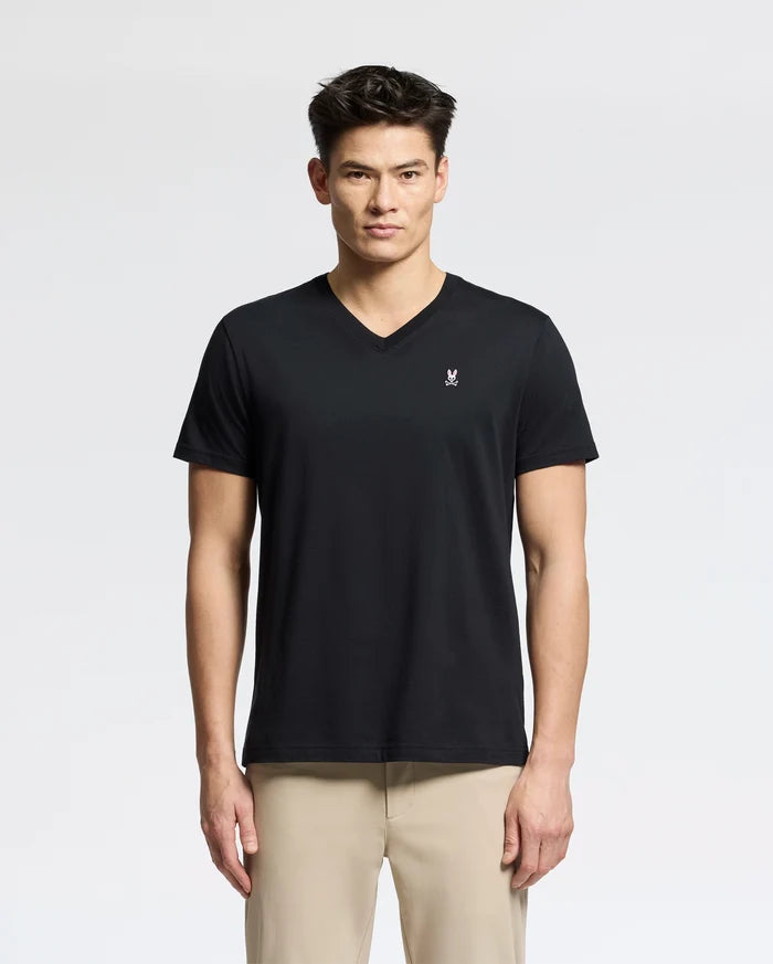 A person in the Psycho Bunny MENS CLASSIC V NECK TEE - B6U100ARPC, made of soft Pima cotton and featuring a small white chest logo, pairs it with light beige pants against a plain white background. They have a neutral expression while wearing this black tee.