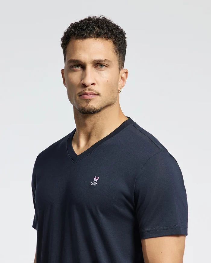 A person with short curly hair and a goatee is wearing the MENS CLASSIC V NECK TEE - B6U100ARPC, a Pima cotton V-neck T-shirt by Psycho Bunny, featuring an embroidered bunny logo in classic navy. The background is plain white.