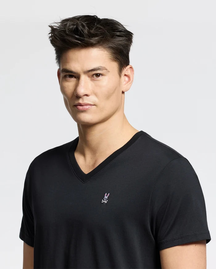 Someone wears a Psycho Bunny MENS CLASSIC V NECK TEE - B6U100ARPC in black, made from soft Pima cotton, with a small logo featuring a rabbit with crossed bones on the chest. The background is plain gray.