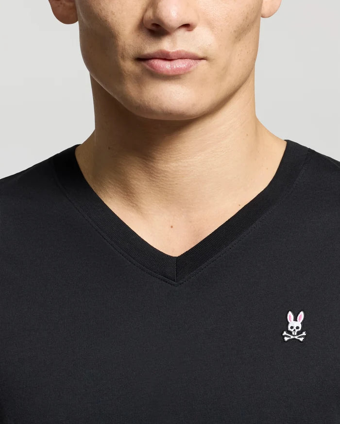 A man wears a Psycho Bunny MENS CLASSIC V NECK TEE (B6U100ARPC), made from premium Pima cotton, featuring a pink bunny head and white eyes above bold crossbones on a light gray background.