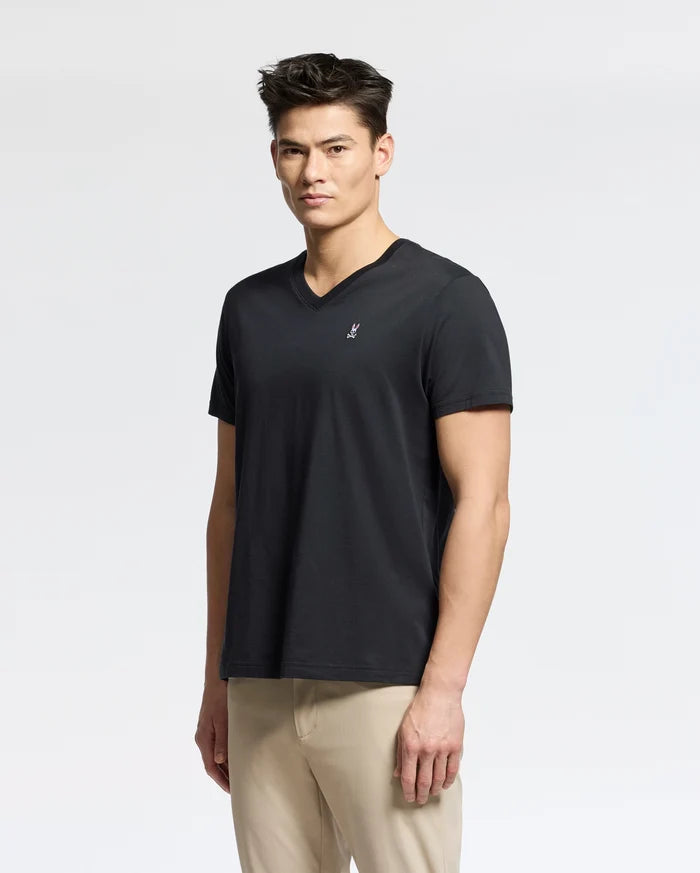 A person faces the camera wearing a black Psycho Bunny MENS CLASSIC V NECK TEE - B6U100ARPC in Pima cotton with a small white embroidered logo on the chest and beige pants, set against a plain white background.