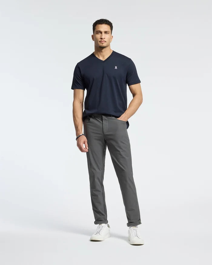 A man stands against a plain background, wearing the Psycho Bunny MENS CLASSIC V NECK TEE (B6U100ARPC) in dark blue, crafted from soft Pima cotton. He pairs it with gray pants and white sneakers. With short curly hair and a relaxed posture, he looks directly at the camera.