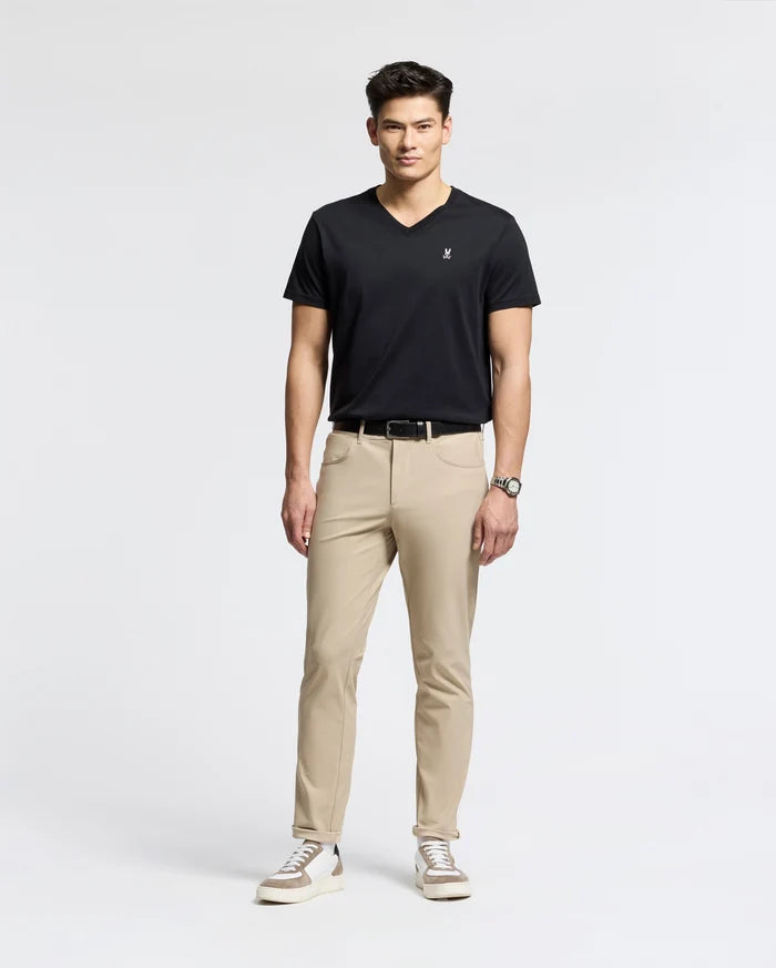 Against a white backdrop, a man models effortless style wearing Psycho Bunny's MENS CLASSIC V NECK TEE (B6U100ARPC) in black. He pairs it with beige pants, white sneakers with black details, and a wristwatch. With short dark hair and relaxed posture, he exudes laid-back elegance.