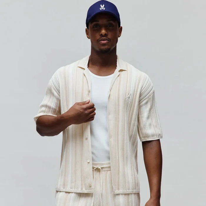 Mens Cliff Shirt, off-white relaxed fit short-sleeve button-up with beige vertical stripes, ribbed knit texture, and small embroidered Psycho Bunny skull logo on left chest. Matching shorts and navy cap with white bunny skull.