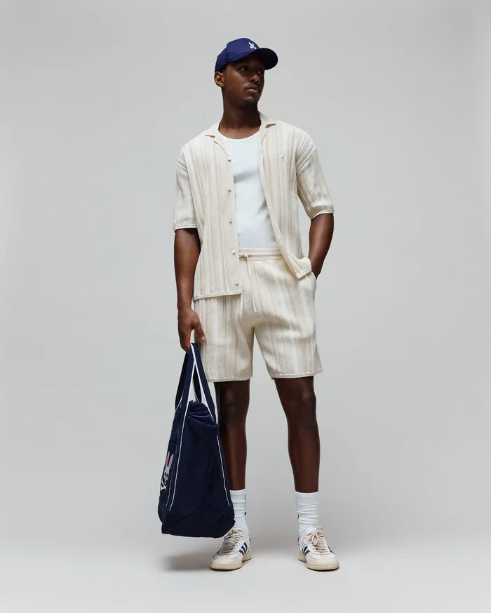 Mens Cliff Shirt, a cream ribbed shirt with small embroidered Psycho Bunny logo, matching relaxed drawstring shorts, white tee, navy cap with white logo, and navy tote with large Psycho Bunny graphic.