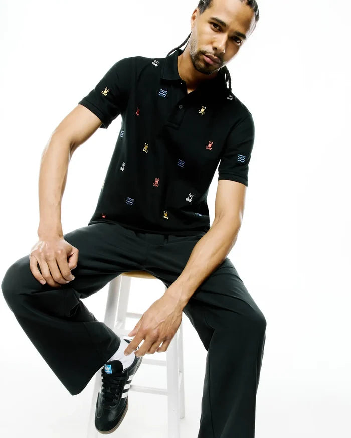 Mens Colin All-Over Pique Polo in black, featuring a classic fit and an all-over embroidered pattern of small multicolor Psycho Bunny skull logos and blue wave graphics.