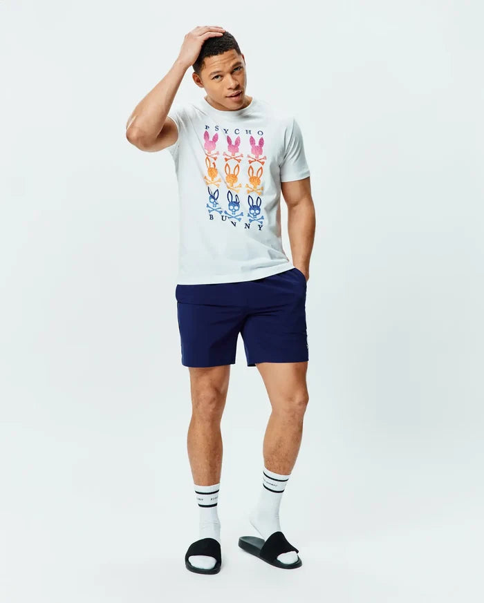 White Mens Colt Graphic Tee, classic fit, soft jersey, with a large central graphic of nine gradient multi-color Psycho Bunny skull logos and text, paired with navy athletic shorts featuring a small white Psycho Bunny logo.