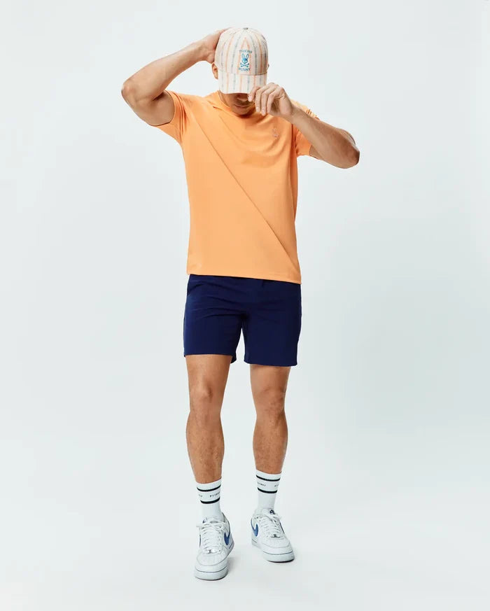 Mens Dash Bunny Sport Tee in peach performance jersey, classic fit, with a small embroidered Psycho Bunny logo on the chest, paired with navy athletic shorts, a striped cap, and white Psycho Bunny socks.