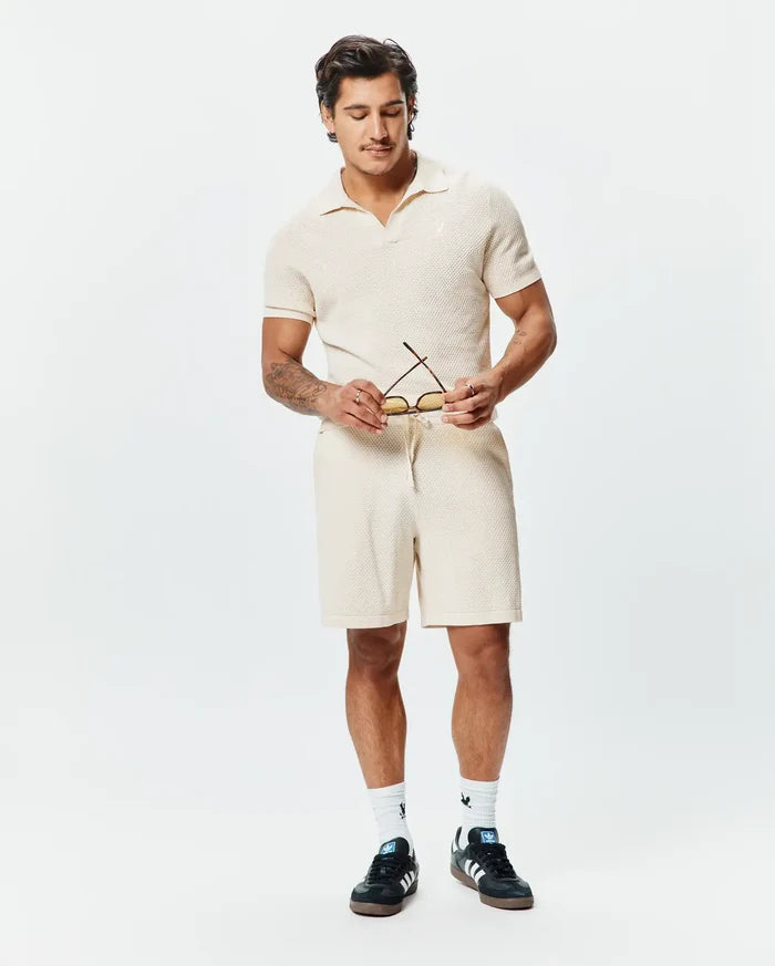 Cream Mens Devon Knit Polo, featuring a classic fit, textured waffle knit fabric, open resort collar, and a small embroidered Psycho Bunny skull logo on the left chest, complemented by matching knit shorts.