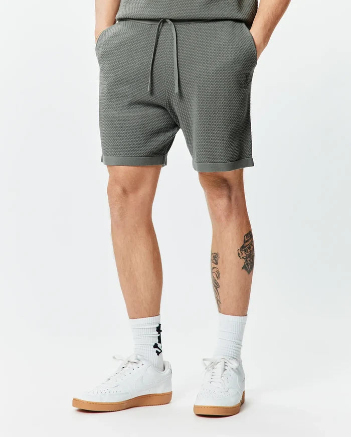 Mens Devon Knit Short 7 in dark olive green, featuring a textured knit fabric, drawstring waist, and a small embroidered Psycho Bunny skull logo on the left thigh, with a relaxed fit.