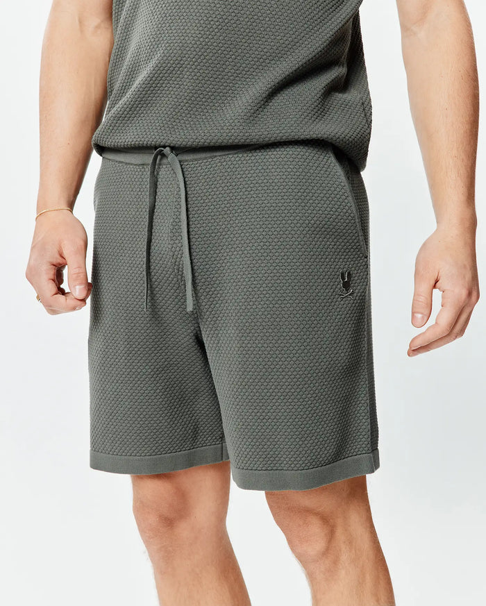 Mens Devon Knit Short 7, a dark olive green textured knit short with a relaxed fit, drawstring waist, ribbed hem, and a small embroidered black Psycho Bunny skull logo on the left thigh.