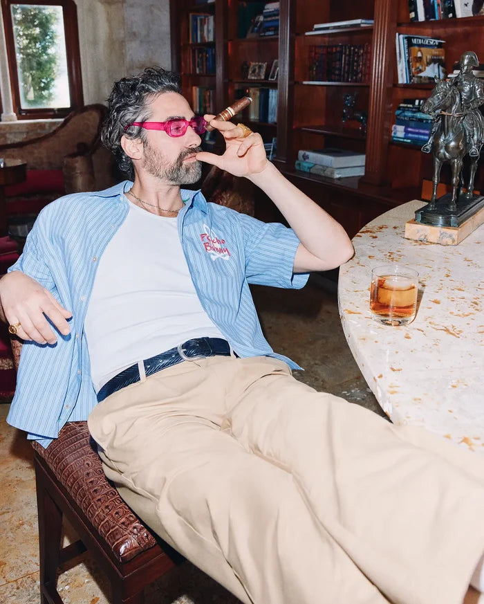 Mens Duncan Stripe Shirt, a light blue short-sleeve button-up with thin white stripes, relaxed fit, and a pink embroidered Psycho Bunny logo on the chest, paired with a white ribbed t-shirt and khaki trousers.