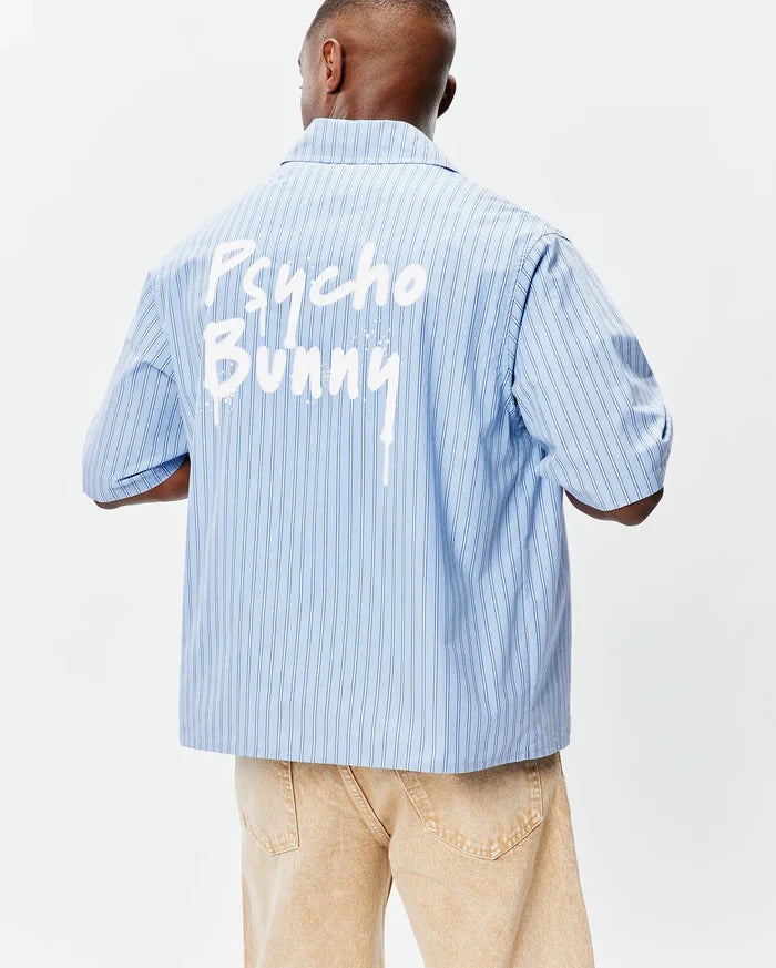 Mens Duncan Stripe Shirt: Light blue pinstripe short-sleeve shirt with a relaxed fit, smooth woven fabric, and a bold white paint-drip Psycho Bunny graphic across the upper back.