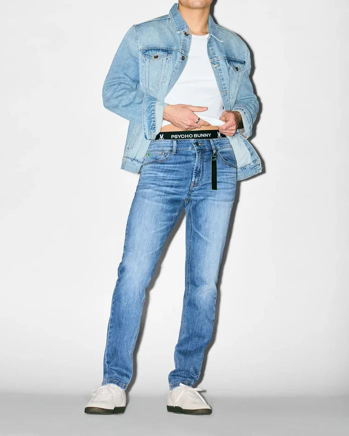 Light blue classic fit denim jacket over a white jersey t-shirt, paired with MENS EMERSON DENIM PANT, a slim fit medium wash denim with subtle fading. Black Psycho Bunny logo waistband with repeating bunny skulls.