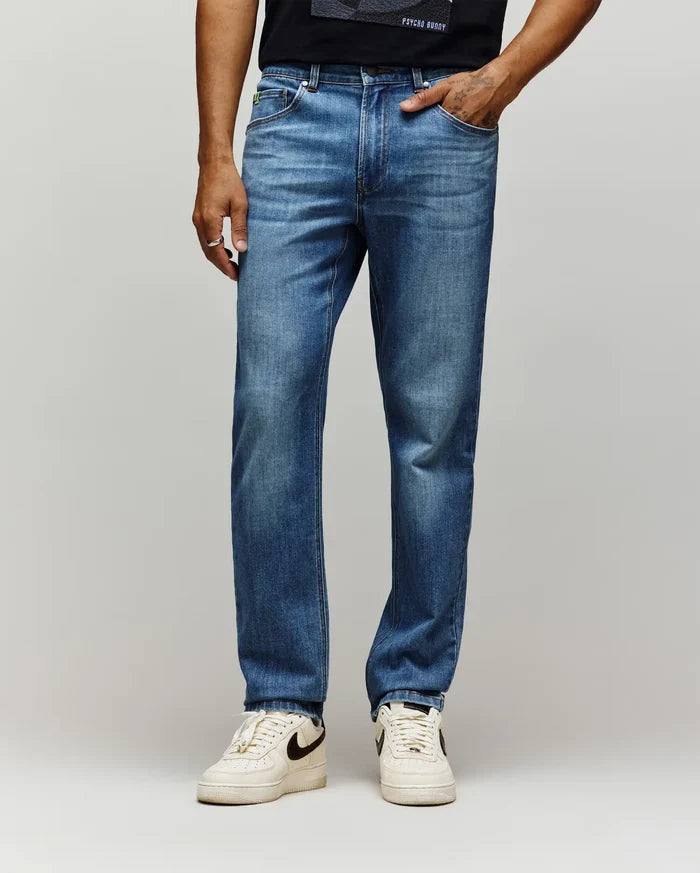 Mens Emerson Denim Pant, classic fit, medium wash blue denim with subtle fading, featuring a small green embroidered Psycho Bunny skull logo on the coin pocket.