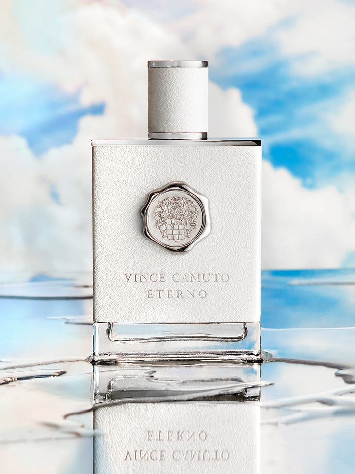 A bottle of Vince Camuto Men’s Eterno 3.4 oz Eau de Toilette Spray stands on a reflective surface, set against a dreamy blue sky with clouds, perfectly capturing the essence of this refined and sophisticated men’s fragrance.