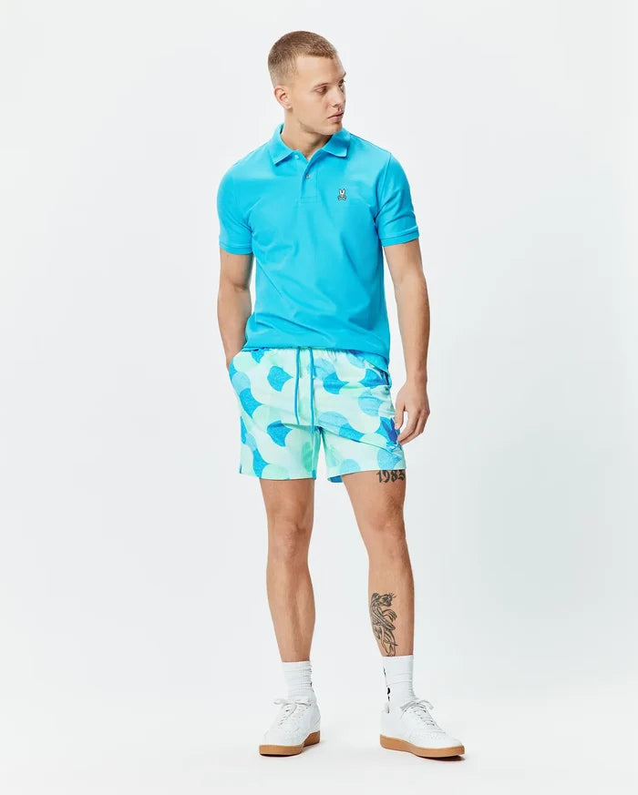 Bright blue pique polo with small embroidered Psycho Bunny skull logo on chest, paired with Mens Ethan Swim Trunk 6 featuring a light mint and blue abstract pattern and drawstring waist.