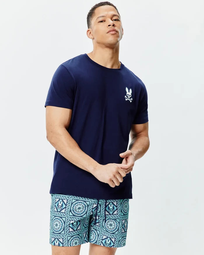 Mens Frank Back Graphic Tee, a classic fit navy blue jersey t-shirt with a small white embroidered Psycho Bunny skull logo on the left chest, paired with teal and blue patterned shorts featuring integrated bunny skull motifs.