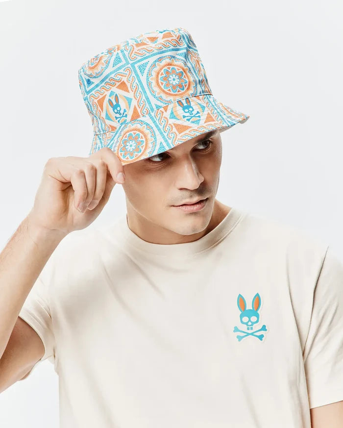 Cream jersey t-shirt with a blue and orange Psycho Bunny skull logo on the left chest, paired with a Mens Frank Reversible Bucket Hat featuring a white, blue, and orange intricate pattern with small bunny skulls.