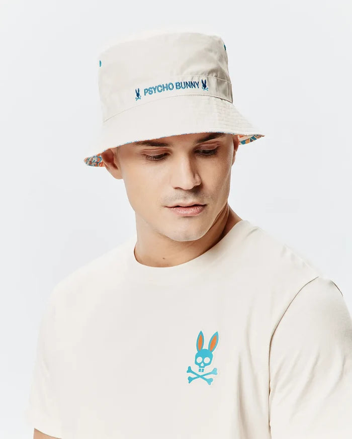 Mens Frank Reversible Bucket Hat in beige with blue Psycho Bunny embroidery and a vibrant patterned underside, paired with an off-white jersey t-shirt featuring a large teal and orange Psycho Bunny skull logo on the left chest.