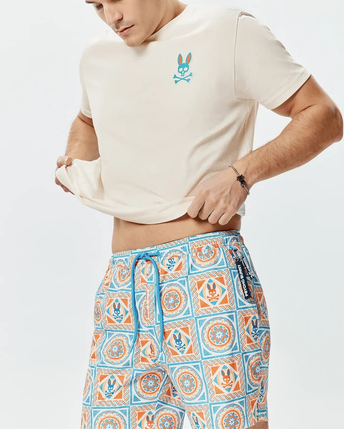Cream jersey t-shirt with a small light blue and orange Psycho Bunny logo on the chest, and Mens Frank Swim Trunk 6 featuring a vibrant orange and blue mosaic pattern with integrated Psycho Bunny skulls and a blue drawstring.
