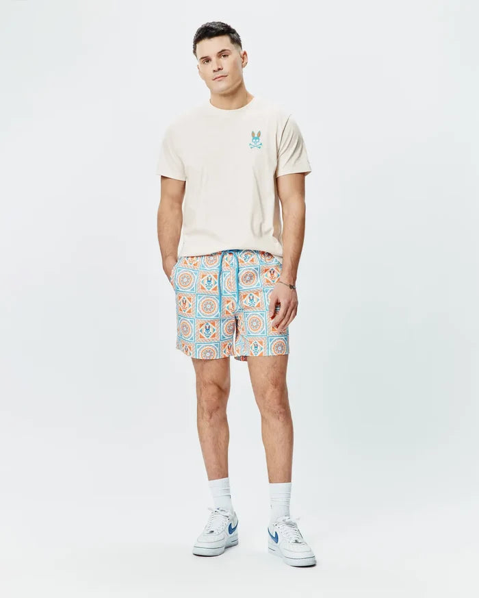 Cream jersey t-shirt with small embroidered blue-orange Psycho Bunny logo on chest, paired with Mens Frank Swim Trunk 6, featuring a vibrant blue and orange geometric tile pattern with integrated bunny skull motifs.