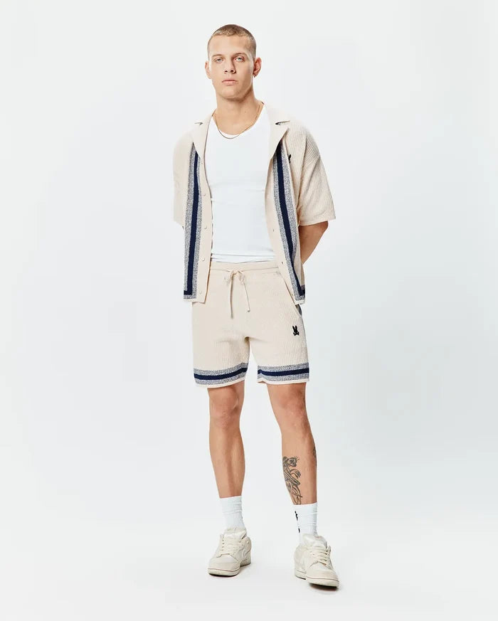 Mens Giles Knit Short 7, a relaxed fit, light beige textured knit short with navy and white striped hem, featuring a small embroidered black Psycho Bunny skull logo on the left thigh and a drawstring waist.