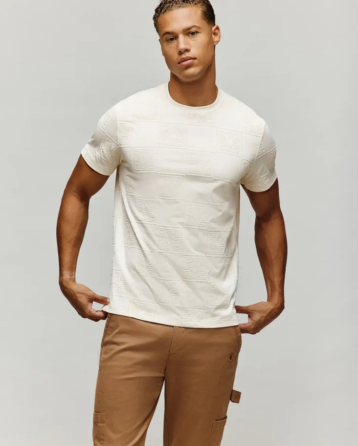 Mens Gio Tee in cream, featuring a classic fit and an all-over embossed checkerboard pattern with Psycho Bunny skull logos and text on a soft jersey fabric.