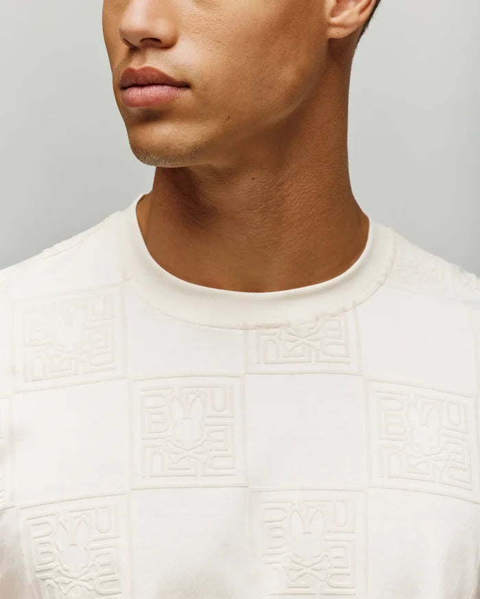 Mens Gio Tee in cream, featuring a classic fit and soft jersey fabric with an all-over embossed checkerboard pattern of the Psycho Bunny skull logo and brand name.