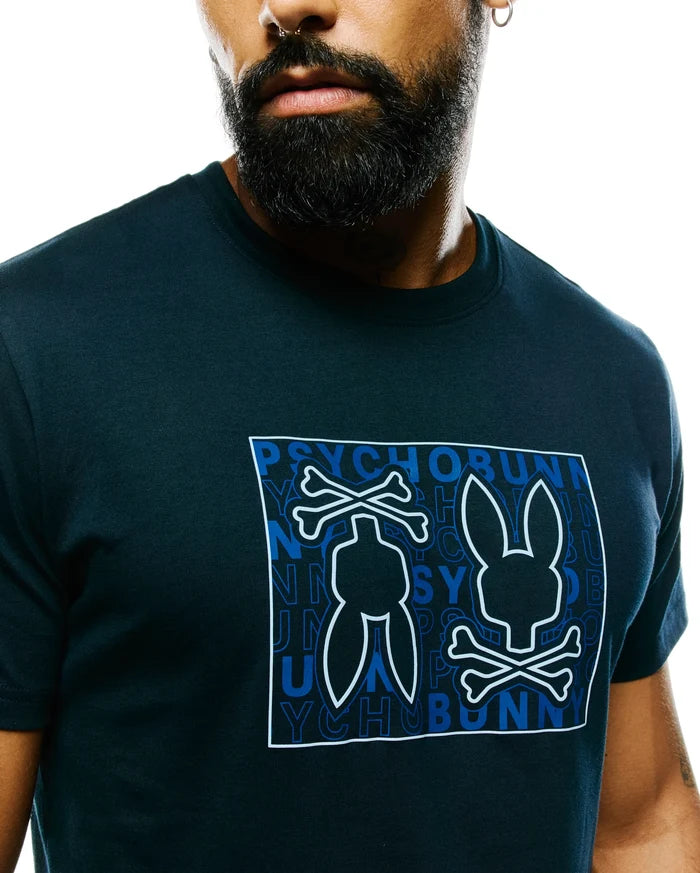 Mens Gold Coast Pima Cotton Graphic Tee: Dark navy jersey t-shirt with a large central graphic featuring the white-outlined Psycho Bunny skull logo over a blue repeated text pattern in a white rectangle, classic fit.