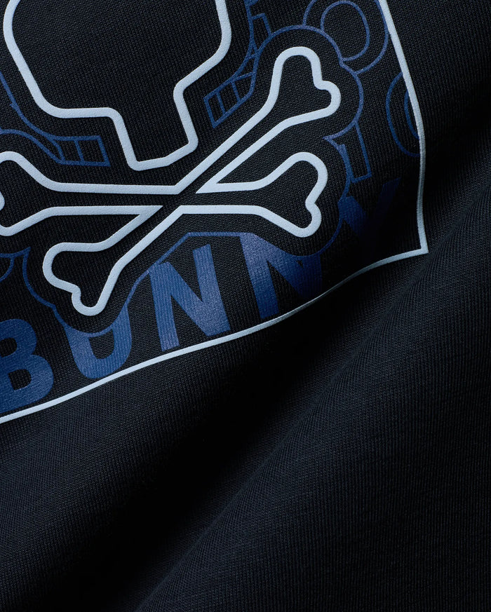 Mens Gold Coast Pima Cotton Graphic Tee in dark navy, featuring a large light blue outlined Psycho Bunny skull and crossbones logo with dark blue geometric patterns and BUNNY text on soft Pima cotton jersey.