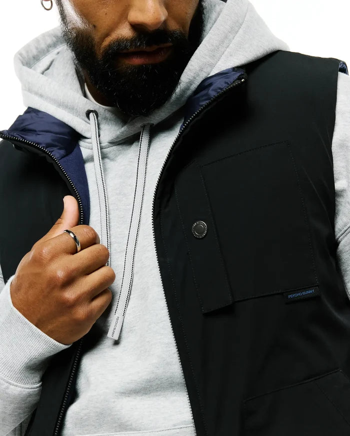 Mens Gordler Reversible Vest in black, smooth fabric, zippered front, snap pocket with blue Psycho Bunny text, navy lining, layered over a grey hoodie with branded drawstrings.
