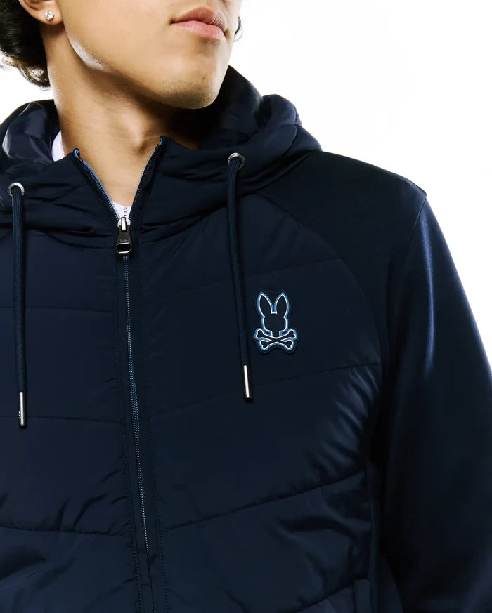 Mens Gordley Mix Media Jacket. Navy blue hooded jacket with a quilted front, knit sleeves, full zip, silver-tipped drawstrings, and an embroidered light blue Psycho Bunny skull logo on the left chest.