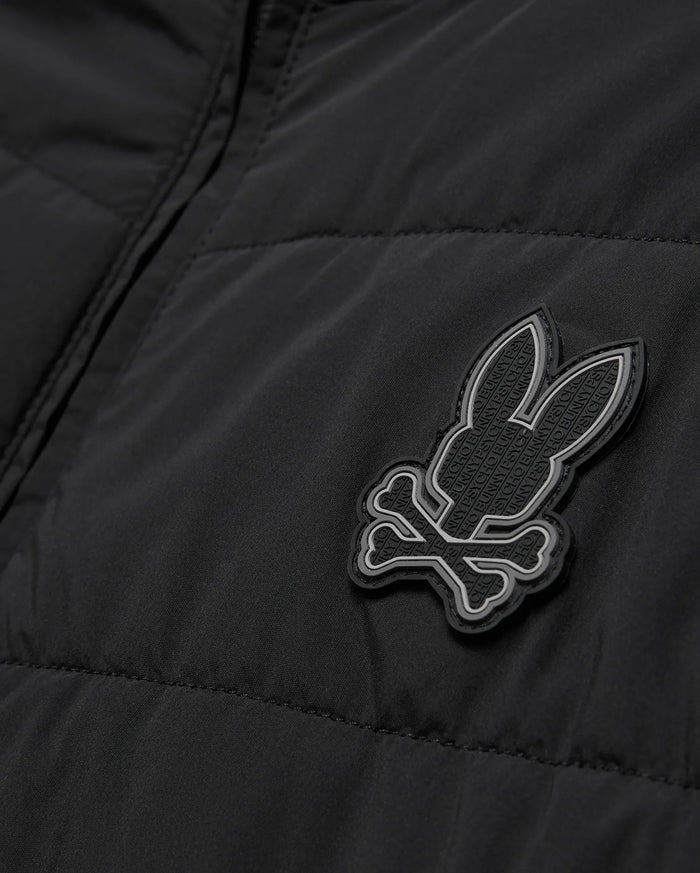 Mens Gordley Mix Media Jacket, black quilted outerwear with a prominent grey and white Psycho Bunny skull logo on the chest, featuring subtle repeating brand text within its design.