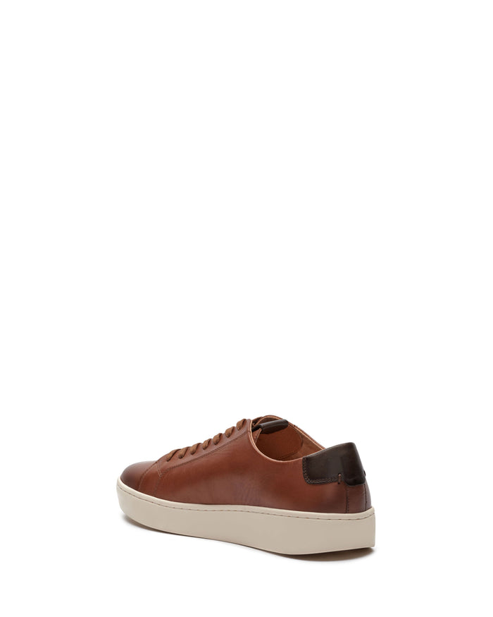 A men’s Hallman casual sneaker by Vince Camuto, featuring brown leather with a white sole and dark brown heel detail, is displayed at an angle with the heel facing forward and to the right on a plain white background, combining stylish design with everyday comfort.