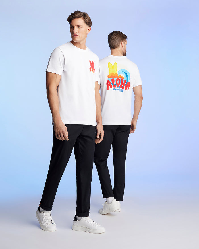 Two men are standing against a light blue background, wearing matching white t-shirts and black pants. The man on the left faces the camera, showcasing a red graphic pattern on his Pima cotton jersey. The man on the right is turned away, revealing a colorful "ALOHA" Hawaii tee design on the back of his MENS HAWAII CITY TEE - B6U169Y1PC by Psycho Bunny.