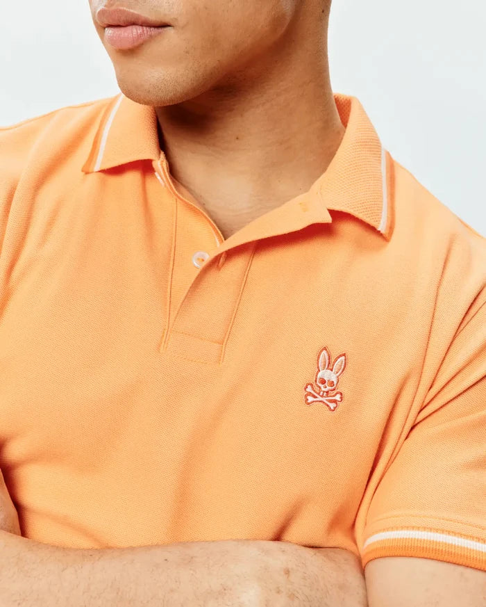 Mens Holden Pique Polo in vibrant orange, featuring a classic fit, pique knit texture, white tipped ribbed collar and cuffs, and an embroidered Psycho Bunny skull logo on the left chest.