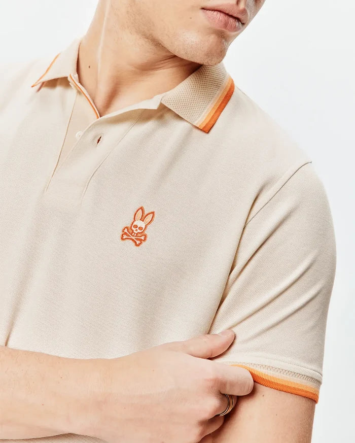 Mens Holden Pique Polo in beige pique knit, featuring a small orange embroidered Psycho Bunny skull logo on the left chest and orange striped collar and sleeve cuffs.
