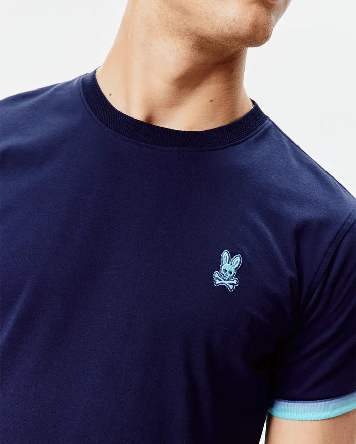 Mens Holden Tee in navy jersey, classic fit, with a small embroidered light blue Psycho Bunny skull logo on the left chest, a ribbed navy crew neck, and striped light blue cuffs.